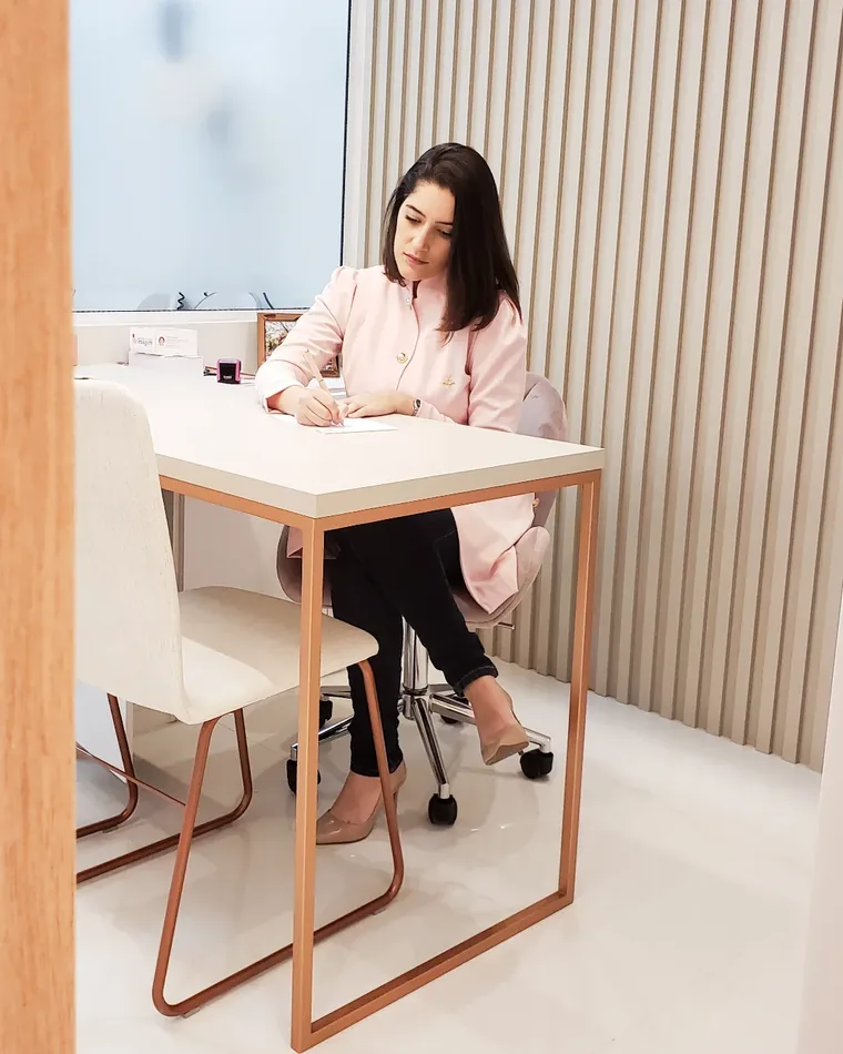 Dra. Ana Vieira de jaleco rosa, sentada à mesa do consultório com parede ripada de madeira